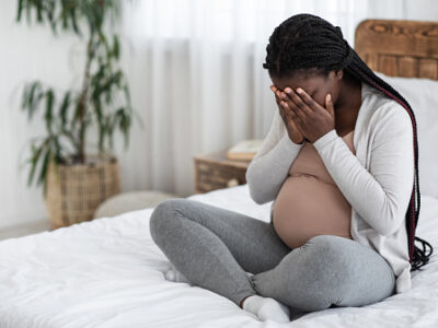 Pregnant Black woman on bed crying, face in hands.